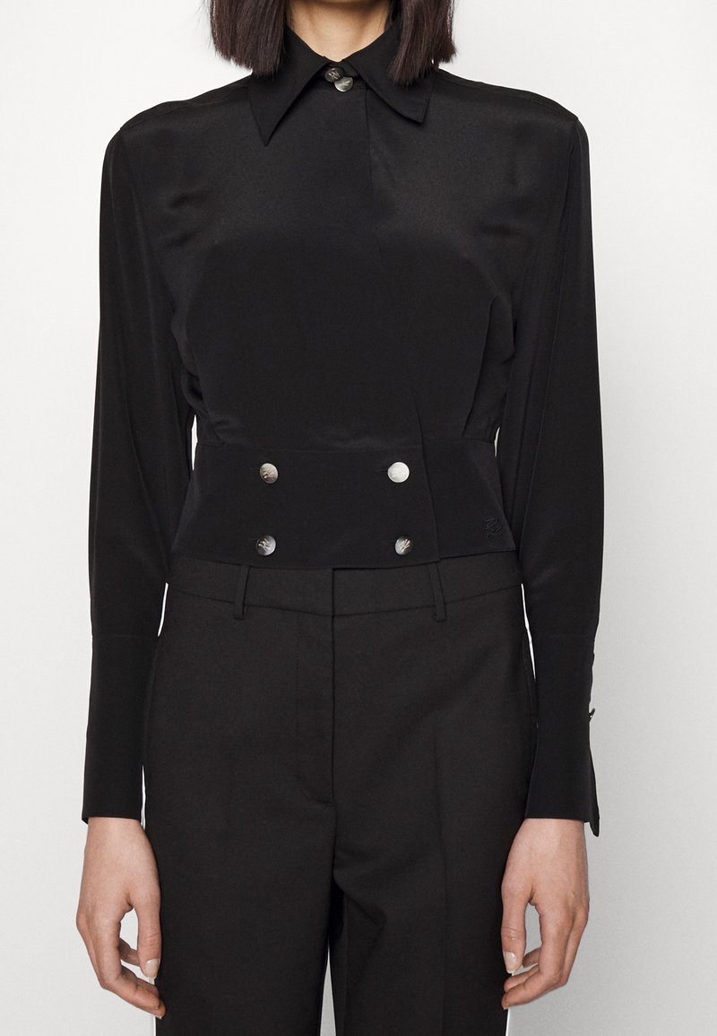 Osoba oblečená v černé košili s dlouhým rukávem a širokým límcem, vybavené čtyřmi stříbrnými knoflíky v pase, doplněná černými přizpůsobenými kalhotami.