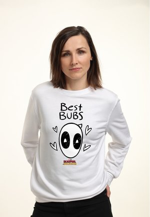 Kvinde med skulderlangt brunt hår iført en hvid sweatshirt med en tegneserie-Deadpool-ansigt, hjerter og teksten "Best BUBS."