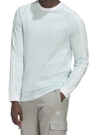 Světle modrá košile s dlouhým rukávem a bílými detaily, s kulatým výstřihem a třemi bílými pruhy na rukávech. Viditelné vyšívané logo.