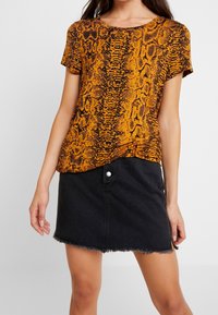 Koszulka z krótkim rękawem w pomarańczowy i czarny wzór w węża, w połączeniu z czarną spódnicą dżinsową z poszarpanym dołem i zapięciem na zatrzask.
