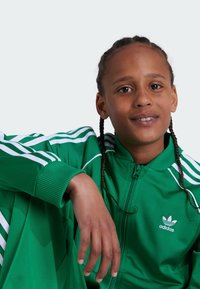 Fato de treino verde em poliéster com uma jaqueta com fecho éclair e riscas brancas, apresentando um logótipo da Adidas bordado no peito. Tecido texturizado.