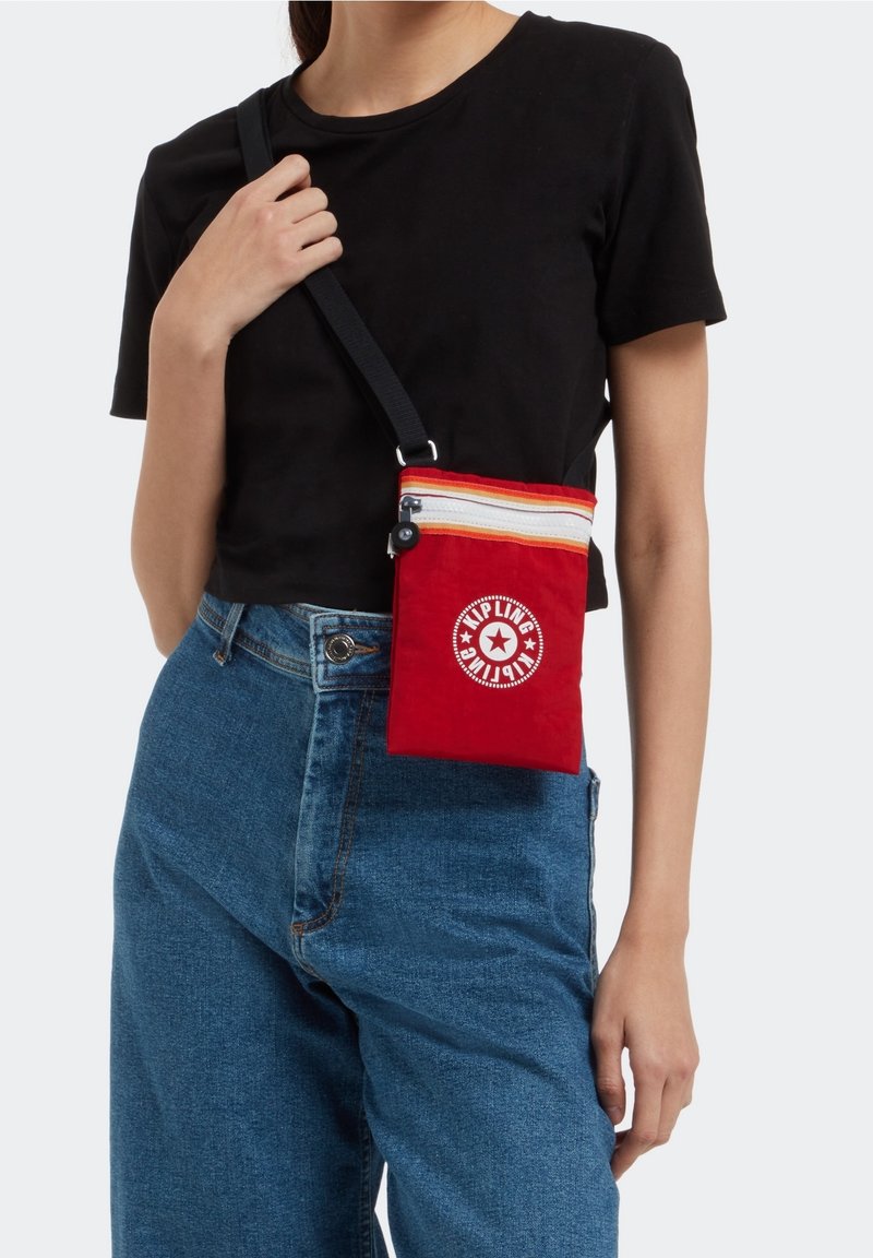 Personne portant un t-shirt noir et un jean bleu porte un petit sac bandoulière rouge avec un logo Kipling blanc et un détail de fermeture éclair rayé.