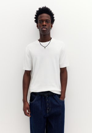 Junger Mann steht vor weißem Hintergrund, trägt ein schlichtes weißes T-Shirt, dunkelblaue Jeans mit Knopfleiste und eine schwarze Perlenkette.