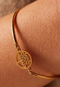 Gouden armband met een ronde bedel met een boomontwerp, rustend op gestructureerde huid. De armband heeft een gladde, gepolijste afwerking.