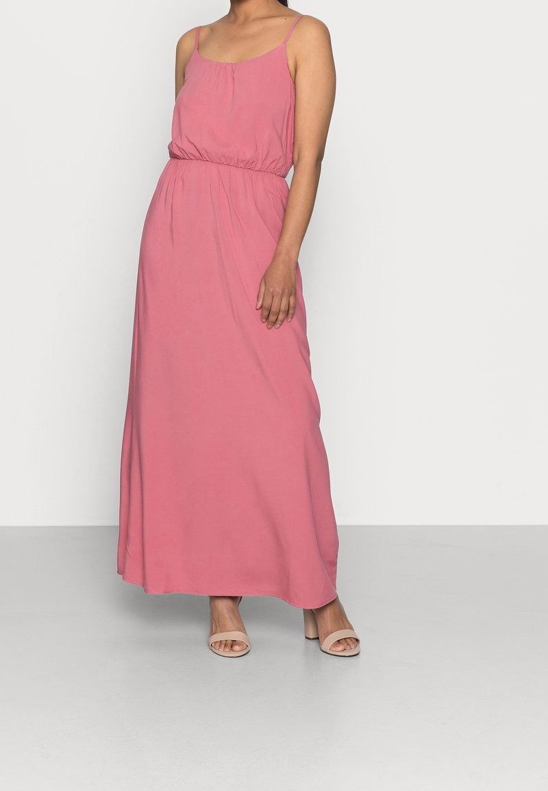 Femme portant une robe maxi rose sans manches avec une taille ajustée et des sandales beiges à talons blocs à bout ouvert, debout sur un fond uni.