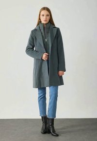Siv šal coat z oblikovano silhueto, zarezanim ovratnikom in stranskimi žepi. Usklajen s jakno na zadrgo, modrimi kavbojkami in črnimi gležnjarji.