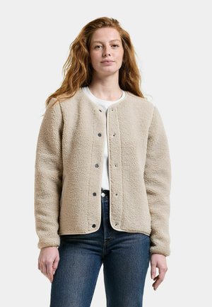 Jeune femme aux longs cheveux roux, portant une veste polaire beige par-dessus une chemise blanche et un jean bleu, debout devant un fond uni.