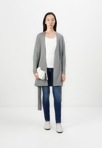 Szara kardiganka z otwartym przodem, wykonana z miękkiego materiału, w połączeniu z białą bluzką i niebieskimi dżinsami. Trzyma kremową torebkę ze sznurkiem.