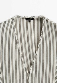 Chemise en lin beige et grise à rayures verticales avec un col en V profond, des bords effilochés et deux petits boutons à l'avant.