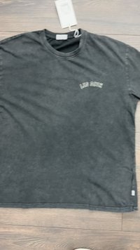 Camiseta negra de cuello redondo colocada plana sobre un suelo de madera, con pequeño texto blanco "LES DEUX" en el pecho y una etiqueta de precio blanca adjunta.