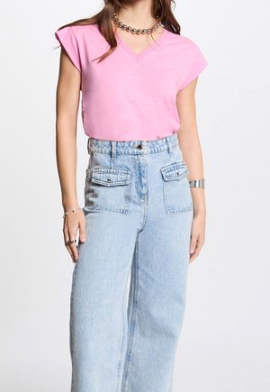 Femme portant un haut rose à manches courtes avec col en V, rentré dans un jean taille haute bleu clair avec poches à rabat devant et des bijoux argentés.