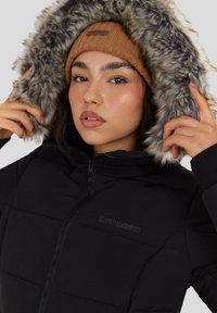 Zwarte gewatteerde jas met een afneembare capuchon voorzien van een faux fur rand. Ritssluiting en gestikte paneelontwerp. Bruine gebreide muts gedragen eronder.