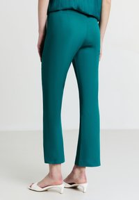 Donna che indossa pantaloni turquoise a lunghezza caviglia e sandali con tacco bianchi, in piedi contro uno sfondo chiaro e uniforme.