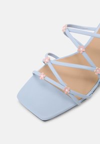 Světle modré sandály na podpatku s úzkými pásky a hranatou špičkou. Na páscích se nacházejí květinové akcenty v růžové a bílé barvě. Hladká imitace kůže.