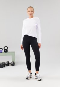Bijela majica dugih rukava i crne tajice napravljene od prozračnog materijala. Outfita se dopunjuje srebrnim i crnim sportskim tenisicama. Vidljiva je i fitness oprema.