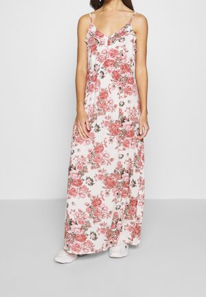 Frau trägt ein weißes Maxikleid mit pinkem Blumenmuster, dünnen Trägern, gerafftem Ausschnitt und weißen Sneakers vor einem schlichten grauen Hintergrund.