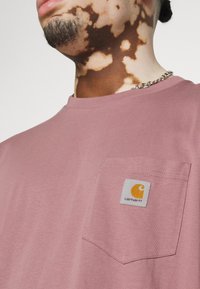 T-shirt en coton rose avec un col rond, poche sur la poitrine gauche, présentant un écusson logo carré en jaune et gris. Texture lisse.