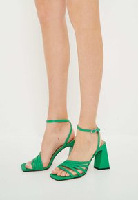 Sandalias de tacón alto verdes con tiras, diseño de punta cuadrada, tiras delgadas en el tobillo y textura de material suave.
