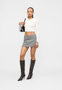 Femme aux cheveux tressés portant un pull court à fermeture éclair blanc, une mini-jupe à carreaux noir et blanc, des bottes noires montant jusqu'aux genoux, et tenant un sac à main noir.