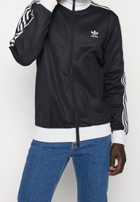 Person som bär en svart Adidas träningsjacka med vita ränder på ärmarna och blå jeans, stående mot en vit bakgrund.