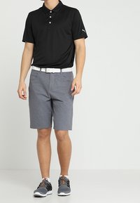 Mies, jolla on musta poolopaita, harmaat shortsit, valkoinen vyö ja harmaat urheilukengät, seisoo yksinkertaisen taustan edessä.