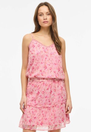 Femme portant une robe sans manches rose à fleurs avec fines bretelles, taille élastique et jupe à volants, debout devant un fond blanc uni.