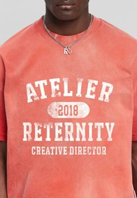 T-shirt couleur rouille avec du texte blanc usé : "ATELIER RETERNITY 2018 DIRECTEUR CRÉATIF." Col rond et manches courtes.