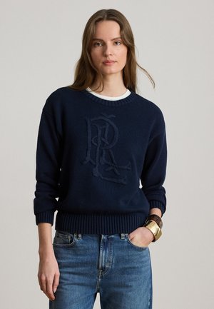 Jeune femme portant un pull bleu marine avec des lettres "RL" texturées, un jean bleu et des bracelets dorés et en écaille de tortue, posant avec une main dans la poche.