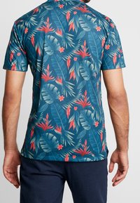 Camisa de manga corta en azul verdoso con un patrón floral tropical. Presenta grandes hojas de palmera y flores rojas, fabricada con una tela suave y liviana.