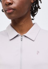 Κοντινό πλάνο ενός ατόμου που φορά ανοιχτό γκρι πουκάμισο με φερμουάρ και γιακά, με ένα μικρό γεωμετρικό λογότυπο στο αριστερό στήθος.