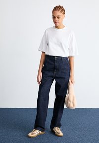 Model wearing a white, oversized short-sleeve shirt, dark blue high-waisted jeans, and brown sneakers, holding a beige bag.