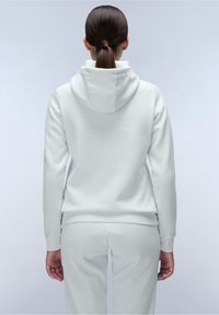 Persona con capelli scuri legati in una coda di cavallo bassa, che indossa un hoodie e pantaloni bianchi, in piedi di spalle rispetto all'osservatore su uno sfondo semplice.