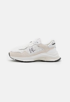 Zapatilla blanca y beige de corte bajo con suela gruesa, cordones en la parte delantera y logotipo negro bordado en el lateral y en la lengüeta del talón.