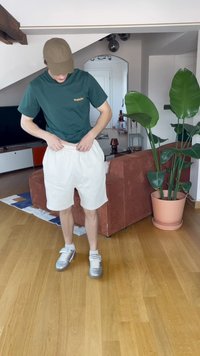 Vihreä lyhythihainen t-paita logolla, valkoiset nyörishortsit ja hopeiset lenkkarit ovat päällä sisätiloissa olevalle henkilölle.