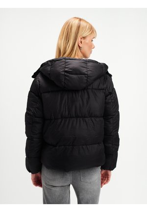 Kobieta z blond włosami, ubrana w czarną kurtkę puchową z kapturem, zwrócona tyłem, oraz jasnoszare dżinsy, na jednolitym tle.