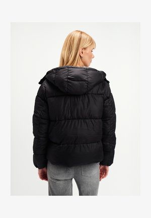 Kobieta z blond włosami, ubrana w czarną kurtkę puchową z kapturem, zwrócona tyłem, oraz jasnoszare dżinsy, na jednolitym tle.