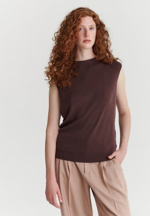 Femme aux longs cheveux roux bouclés porte un haut marron sans manches et un pantalon beige, debout devant un fond clair uni.