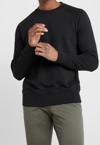 Sudadera negra de cuello redondo con mangas raglán, que presenta puños y dobladillo de canalé, hecha de tejido suave, llevada sobre pantalones verde oliva.