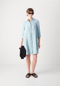 Ανοιχτό μπλε πουκαμίσα-φόρεμα από λινό με κουμπιά μπροστά, μανίκια που μπορούν να αναδιπλωθούν και τσέπη με μπατέ. Συνδυασμένο με μαύρες σανδάλια slide και μαύρο πουλόβερ.
