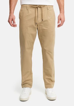 Mann iført beige bukser med snor og hvite joggesko, står mot en nøytral bakgrunn.