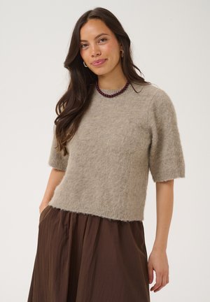 Kvinde med langt brunt hår iført en fuzzy beige kortærmet sweater, brun nederdel, vinrødt perlehalskæde og store øreringe.