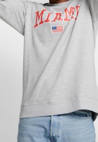 Sweatshirt gris avec "MIAMI" imprimé en rouge accompagné d'une icône du drapeau américain. Le design est décontracté avec un col rond et des poignets côtelés.