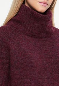 Maglione a collo alto bordeaux realizzato in morbido tessuto lavorato a maglia, con una texture a coste e un colletto alto, che offre un design morbido.