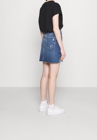 Denimrok met een rafelige zoom, uitgevoerd in een klassieke blauwe wasbeurt, met twee achterzakken en een getailleerde snit. Gekleed met een zwarte shirt en witte sneakers.
