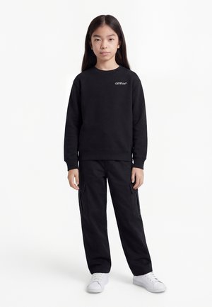 Νεαρός με μακριά σκούρα μαλλιά που φοράει μαύρο φούτερ Off-White, μαύρο παντελόνι cargo και λευκά αθλητικά, στέκεται μπροστά σε λευκό φόντο.