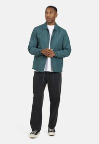 Giacca leggera color teal con design a bottoni, polsini elasticizzati e orlo con coulisse, abbinata a jeans scuri e sneakers nere.