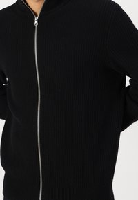 Pull-over zippé noir en tricot côtelé avec un col haut, une fermeture éclair argentée et une coupe ajustée. Tissu texturé sans motifs visibles.