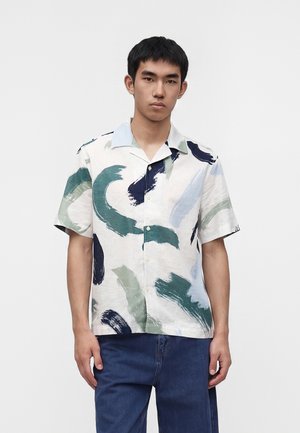Ung mand iført kortærmet skjorte med knapper med grønne, marineblå og blå penselstrøgsmønstre og blå jeans, stående mod en ensfarvet baggrund.