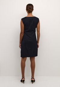 Robe sans manches bleu marine avec des rayures verticales, coupée au genou et avec un dos droit. Portée avec des chaussures à talons hauts noires.