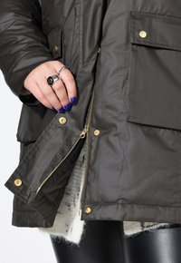Parka isolato di un verde scuro, con dettagli in oro, chiusura a zip e tasche anteriori. Foderato con un tessuto chiaro e strutturato.
